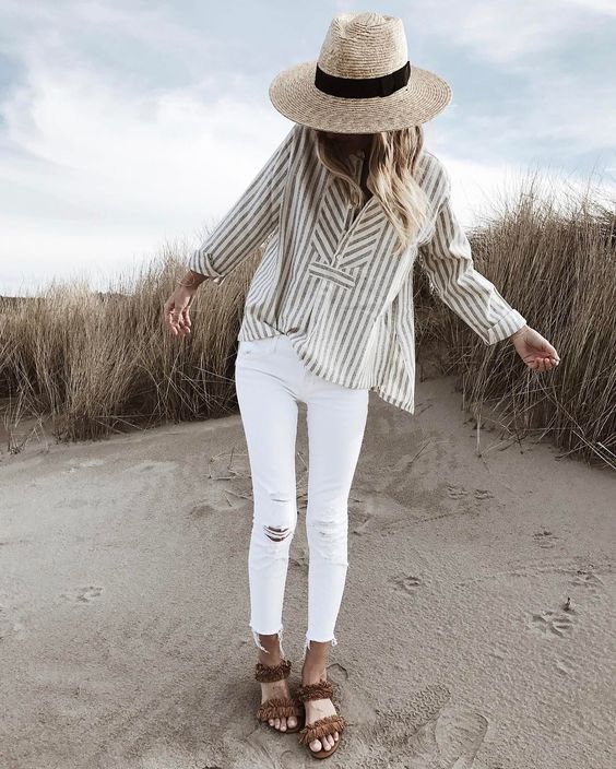 На девушке легкая свободная рубашка в полоску, белые джинсы скинни, босоножки на плоской подошве с бахромой и соломяная шляпа.