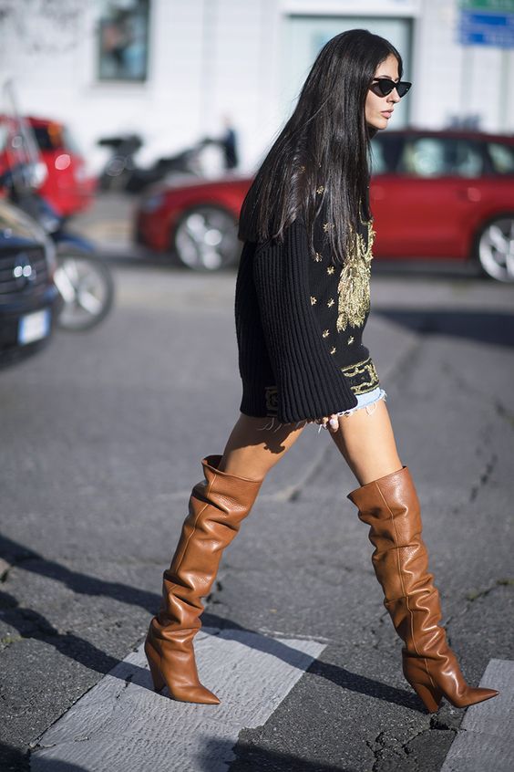На девушке джинсовые шорты, удлиненный черный свитер с широкими рукавами и золотым вышитым рисунком, коричневые кожаные сапоги-”гармошки” с заостренным носом и широким невысоким каблуком, очки.