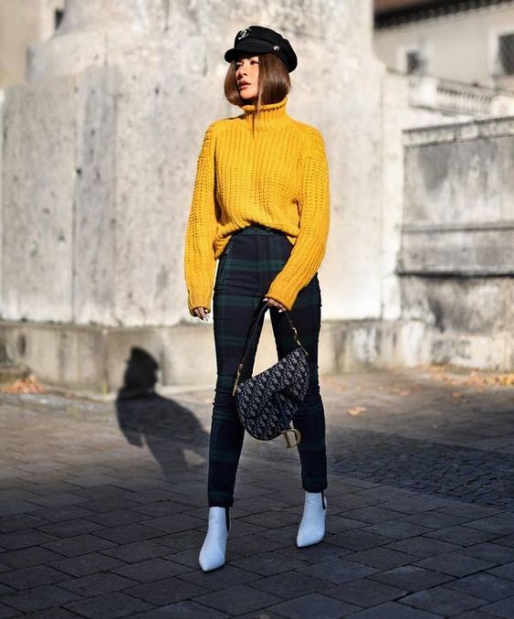 На девушке яркий желтый свитер крупной вязки с объемным горлом, облегающие брюки в крупную клетку, белые ботинки на каблуке и с заостренным носом, сумка асимметричной формы, черное кепи.