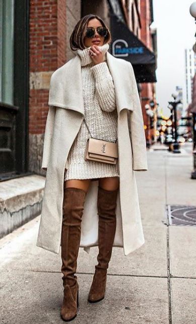 На девушке белый вязаный объемный свитер-платье с горлом, белое удлиненное пальто, замшевые коричневые высокие ботфорты. Образ дополняет маленькая поясная сумка и очки.