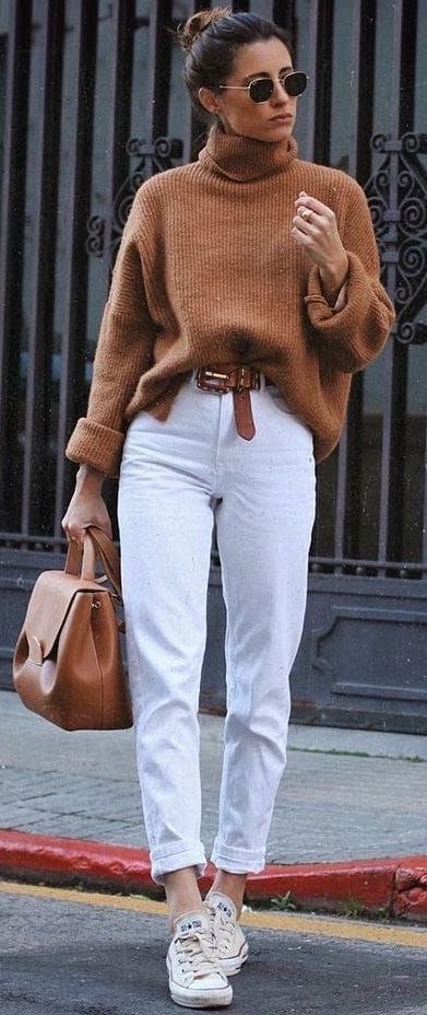 Коричневый свитер с объемным горлом, белые прямые джинсы с коричневым поясом, в сочетании с белыми кедами, коричневой сумкой и черными очками.