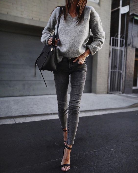 На девушке серый свитер с объемными рукавами, серые джинсы-скинни, черная кожаная сумка и босоножки на каблуке.