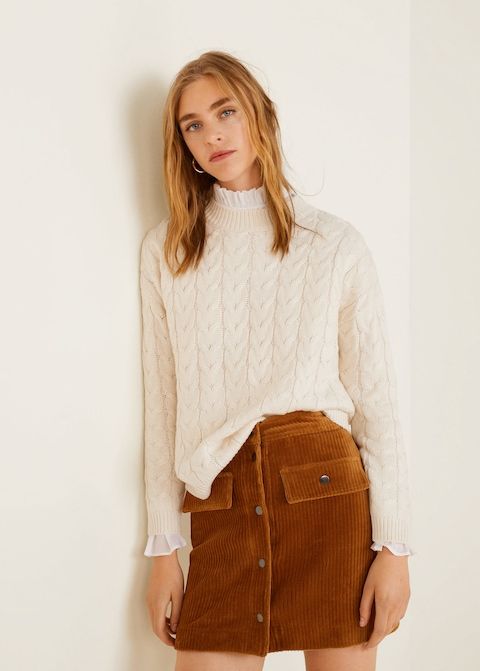 На девушке белый свитер “косичка”, белая рубашка, велюровая прямая коричневая юбка-мини.