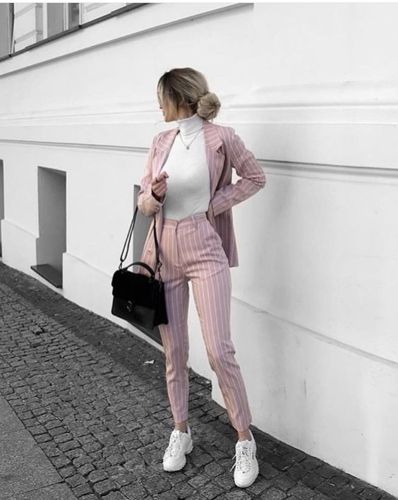 Белая, плотно облегающая, водолазка с воротом в сочетании со светло-розовым костюмом в белую вертикальную полоску: классический удлиненный пиджак и облегающие брюки с высокой талией, белые кроссовки, черная сумка-мессенджер.