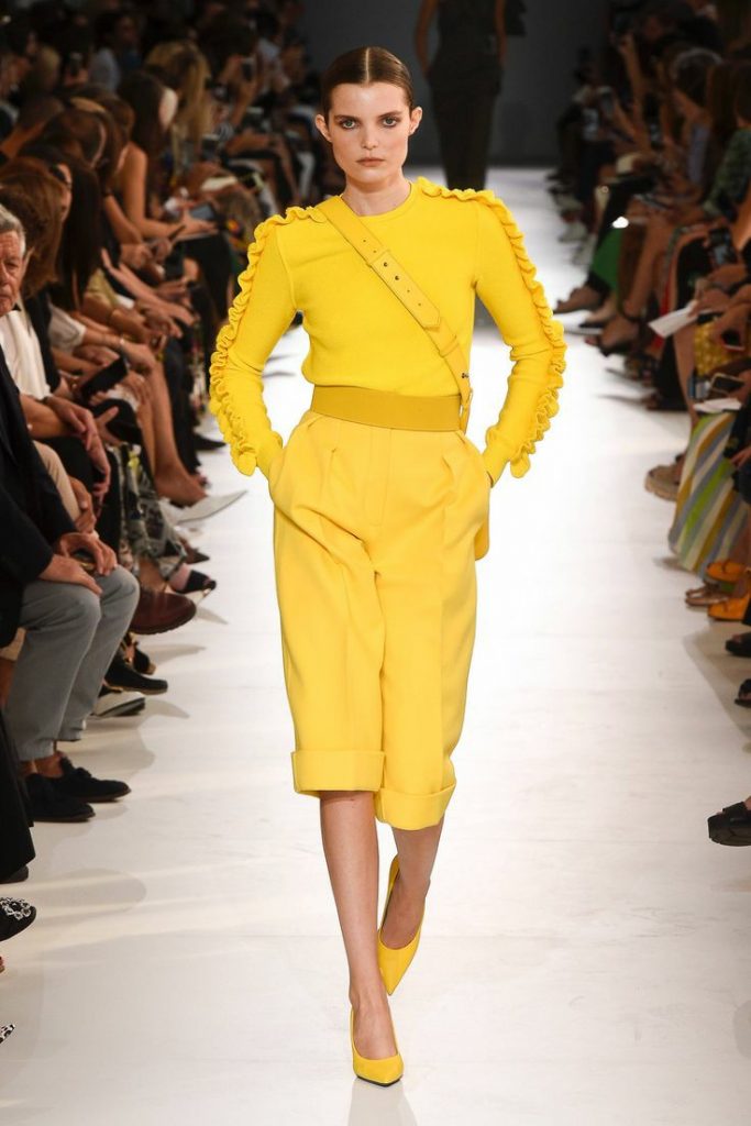 На модели желтая водолазка без ворота, декорированная рюшами на рукавах, желтые свободные шорты-бермуды по колено и с резиновым поясом, желтые лодочки с заостренным носом на каблуке.